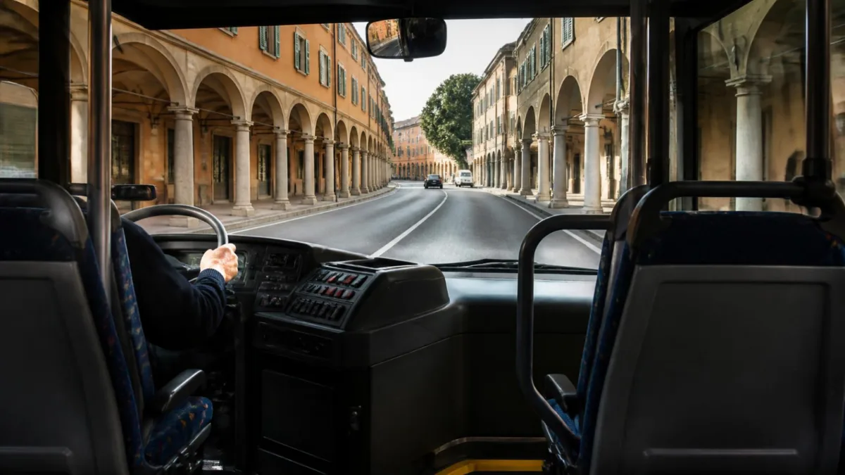 Interno di un autobus visto dal corridoio: mano del conducente sul volante e strada italiana con portici oltre il parabrezza